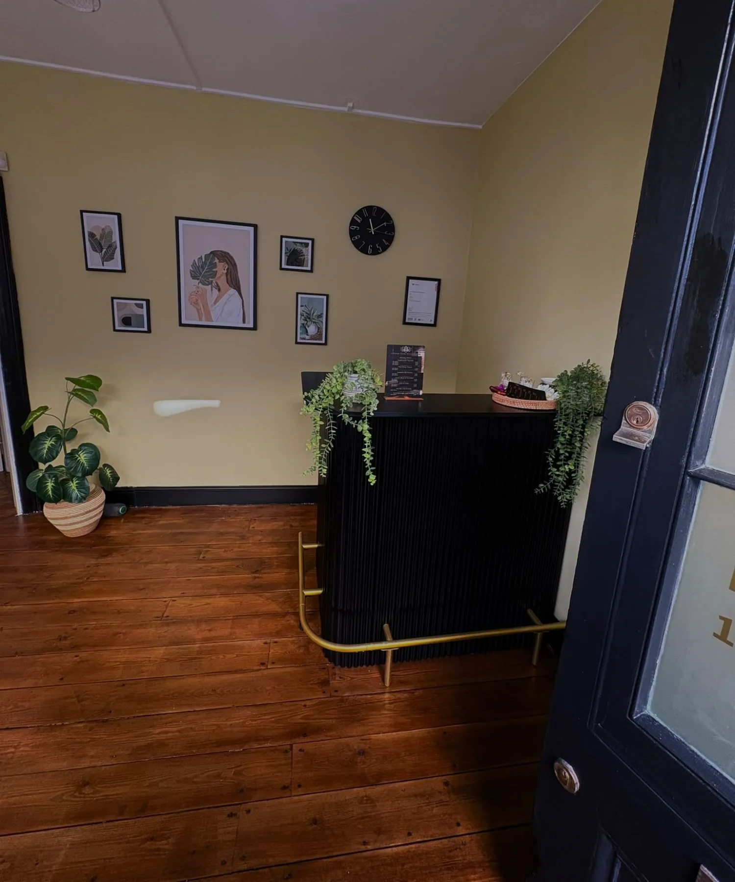 Reception at Lanna, seen from the front door: a black fluted counter with a brass foot rail and trailing plants, framed botanical prints and a black clock on a warm yellow wall, old wooden floorboards